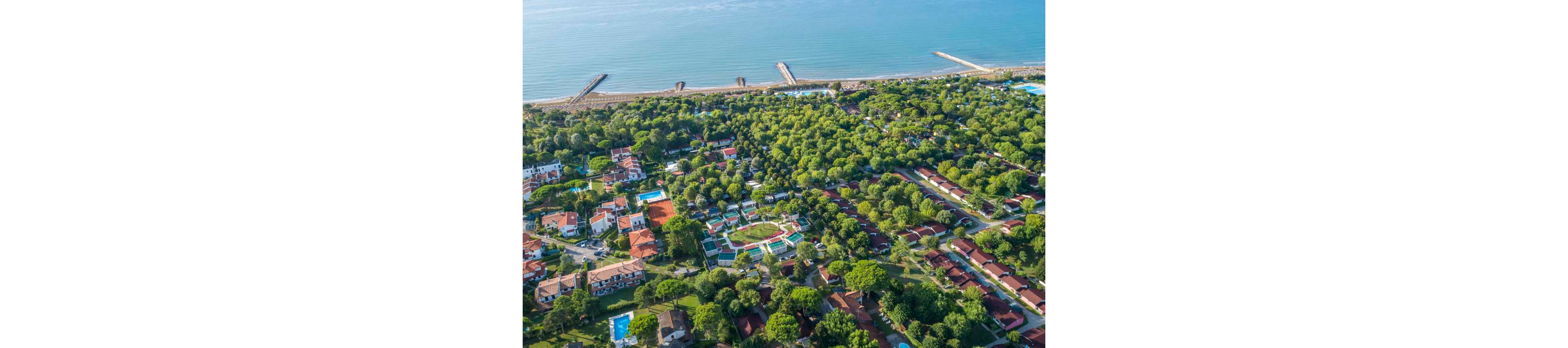 This image shows an aerial perspective of a coastal resort area bordered by the sea. The scene features numerous villas and buildings surrounded by dense green trees, with several swimming pools visible among the properties. The coastline includes sandy beaches and piers extending into the water, creating a tranquil and inviting atmosphere. The overall palette is dominated by greens and blues, evoking a relaxed, vacation mood.