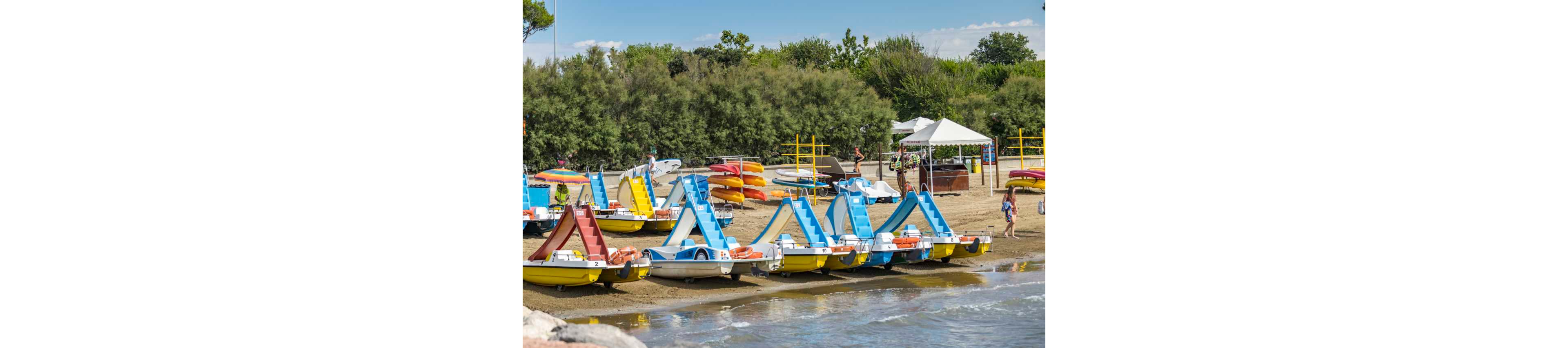 Several brightly colored pedal boats are parked along the shoreline, with their slides facing the water. The scene is set on a sandy beach with kayaks stacked in the background and a white tent providing shade. People, including adults and children, are visible enjoying the outdoor setting, surrounded by lush greenery and clear skies.