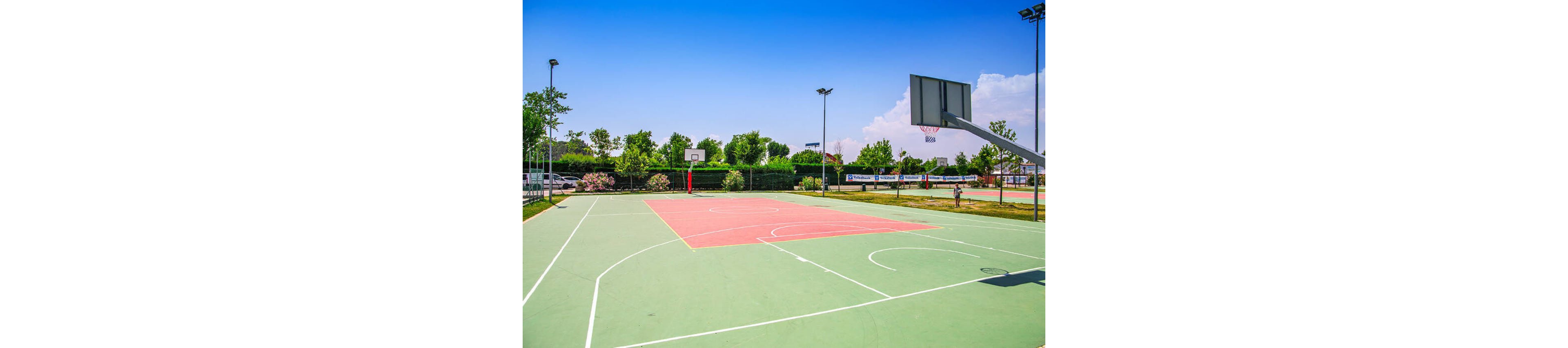 The image features a spacious outdoor basketball court with two hoops, surrounded by greenery and trees. The court is empty, with visible white lines marking the playing area and a red central section. The setting is bright and sunny, with a vivid blue sky and scattered clouds, creating a vibrant and energetic mood. No visible text or numbers are present in the image.