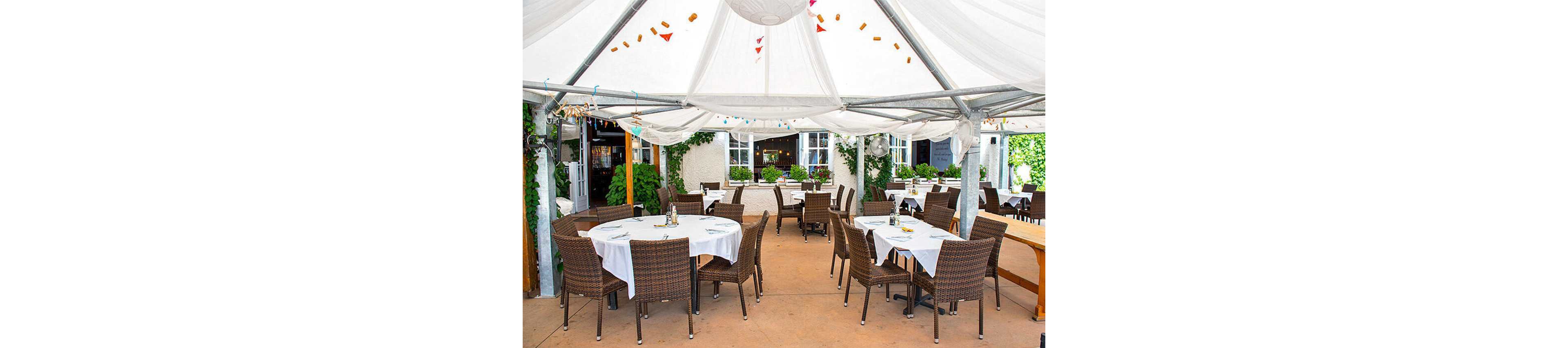 The image shows an outdoor dining area with round tables and wicker chairs arranged under a large white canopy. The setting is bright and inviting, with greenery and potted plants adding a fresh touch. The tables are covered with white tablecloths, and the space appears ready for guests, suggesting a relaxed and elegant atmosphere. No visible text or numbers are present in the image.
