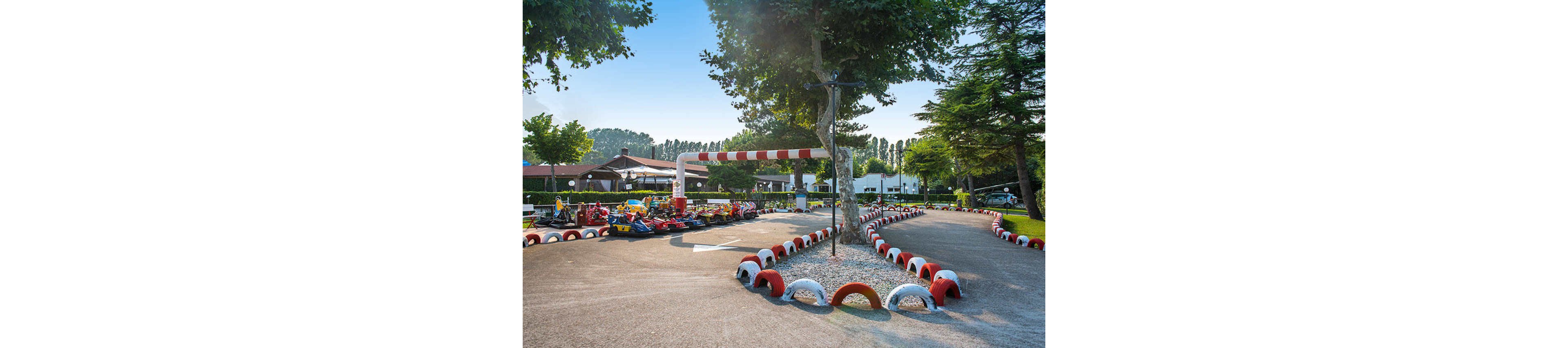 A sunlit outdoor go-kart track features red and white tire barriers lining the winding course. Several go-karts are parked near the starting area, with trees and buildings visible in the background. The setting is calm and recreational, with clear skies and lush greenery enhancing the inviting atmosphere. No visible text or numbers are present in the image.