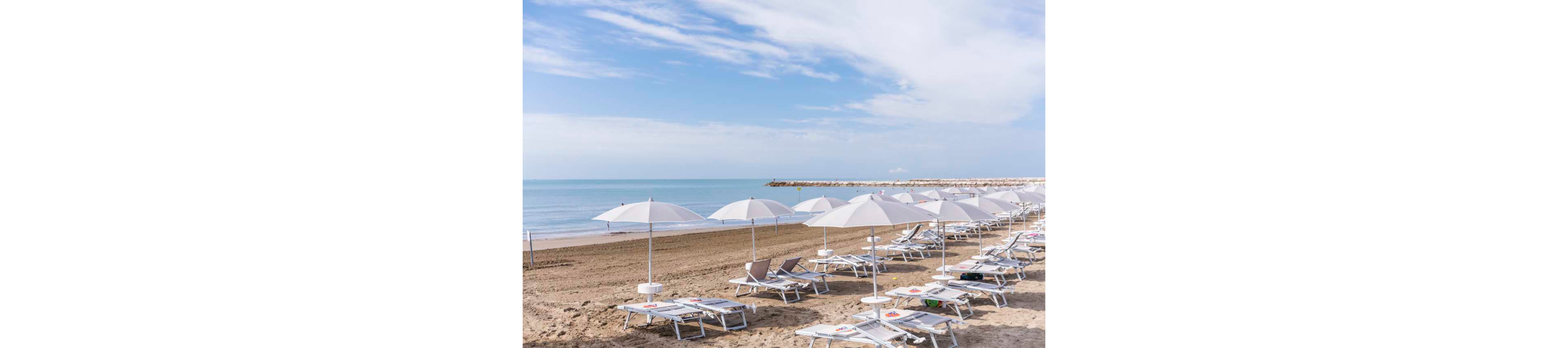 A row of white sun loungers and umbrellas is neatly arranged on a sandy beach facing calm blue water. The scene is tranquil with no people present, suggesting early morning or off-season. The sky is partly cloudy, and the overall palette is soft and serene. The horizon and a distant breakwater are visible, enhancing the peaceful coastal atmosphere.