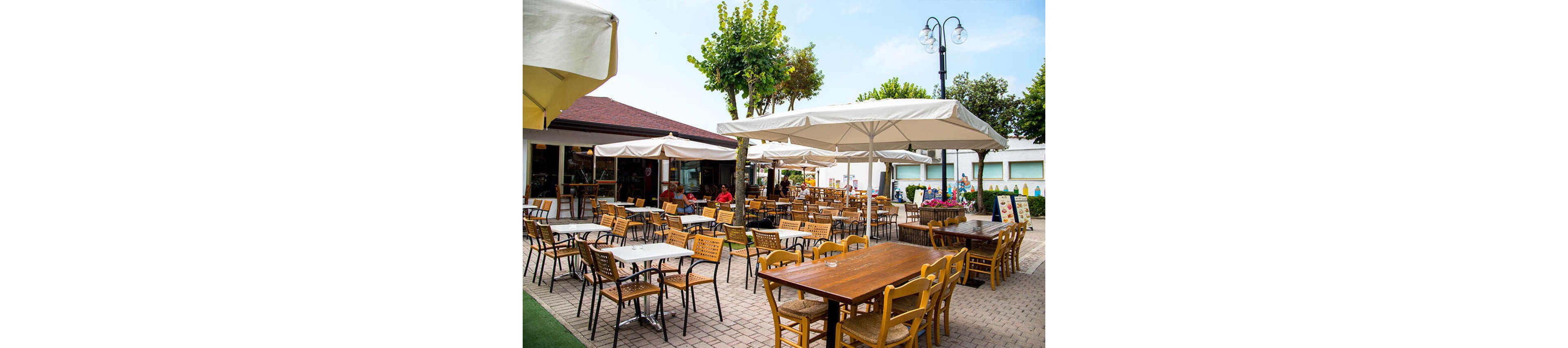 An outdoor café features numerous wooden and metal tables and chairs arranged on a paved patio. Large white umbrellas provide shade, and a few people are seated in the background, enjoying the relaxed atmosphere. The setting is bright and inviting, with green trees and a clear sky adding to the casual, summery mood.