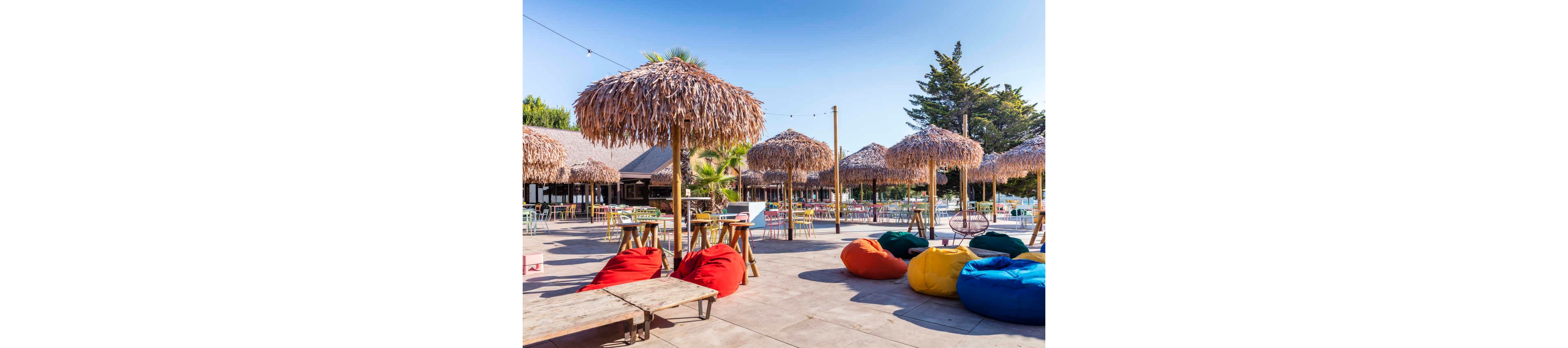 A vibrant outdoor bar area features multiple straw umbrellas and wooden tables, set on a spacious patio. Colorful bean bags in red, yellow, blue, and green provide relaxed seating, while string lights add a festive touch. The setting is bright and sunny, surrounded by palm trees and rustic buildings, evoking a casual, tropical atmosphere.