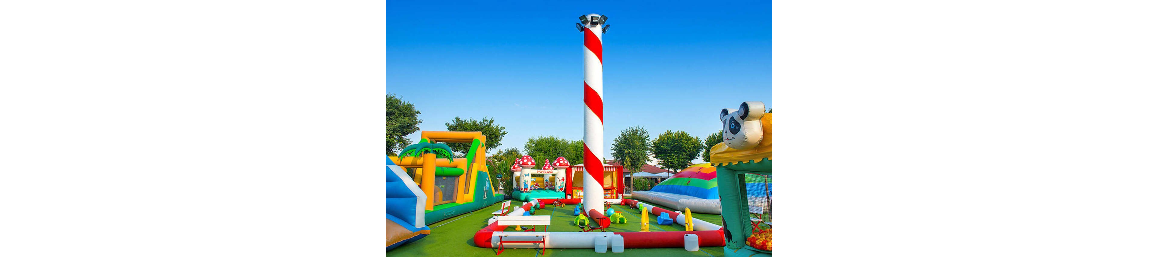 A vibrant outdoor playground features multiple inflatable structures, including slides and bounce houses, set on a green play surface. A tall, red-and-white striped pole stands prominently in the center, surrounded by playful obstacles and benches. The background includes whimsical mushroom-shaped inflatables and a panda-themed structure, all under a clear blue sky. The scene is lively and designed for children’s recreation. No visible text or numbers are present in the image.