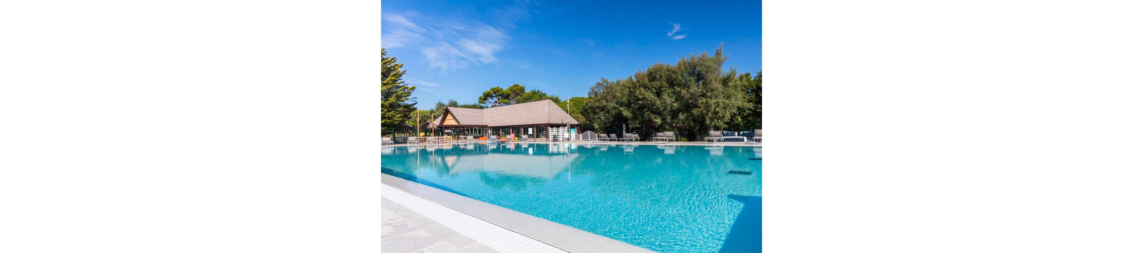A large, clear swimming pool is the main focus, set outdoors under a bright blue sky. The pool is surrounded by lounge chairs and shaded by tall trees, with a resort building visible in the background. The tranquil setting suggests a relaxing vacation atmosphere, with no people present in the scene. The water reflects the sky, enhancing the serene and inviting mood.