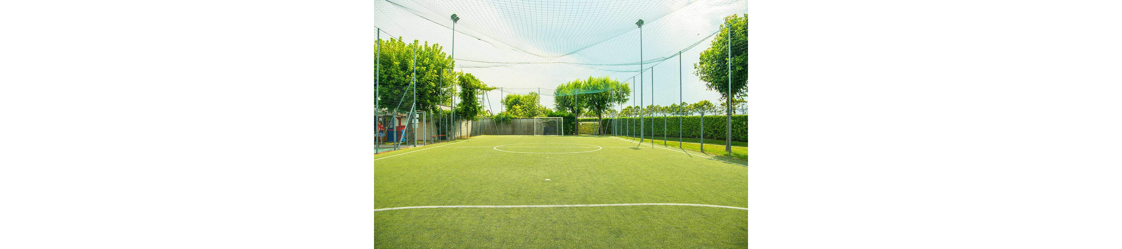 A small outdoor soccer field is shown with bright green artificial turf and a netted enclosure overhead. The field is surrounded by tall fencing and lush trees, creating a vibrant and inviting atmosphere. White field markings are clearly visible, and the area is well-lit by natural daylight. No people are present, emphasizing the peaceful and ready-to-play setting.
