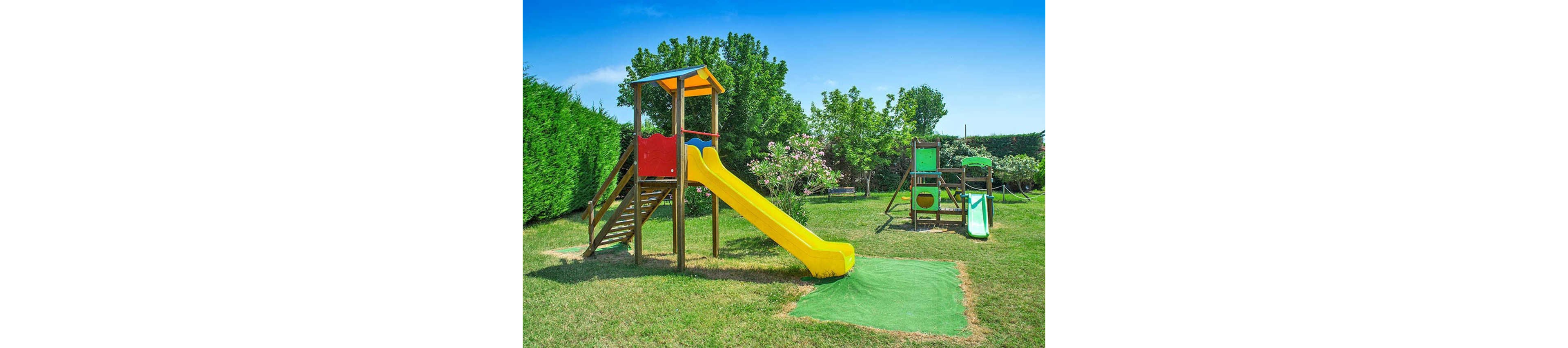 A vibrant children's playground features a yellow slide attached to a wooden climbing structure with a red and blue roof. The play area is set on a grassy lawn surrounded by lush green trees and bushes, with additional equipment including a swing set and a smaller slide. The scene is brightly lit under a clear blue sky, creating a cheerful and inviting atmosphere. No visible text or numbers are present in the image.
