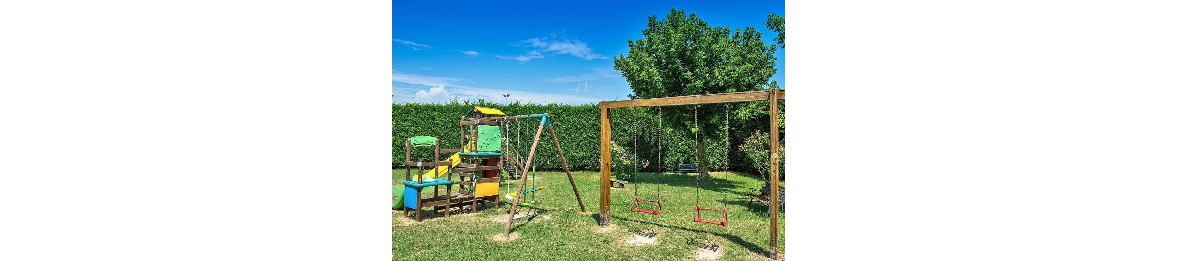 A colorful outdoor playground is set on a grassy lawn under a bright blue sky. The play area features a wooden climbing structure with slides and ladders, alongside a swing set with two red swings. Tall green hedges and a leafy tree provide a natural backdrop, creating a cheerful and inviting atmosphere for children. No visible text or numbers are present in the image.