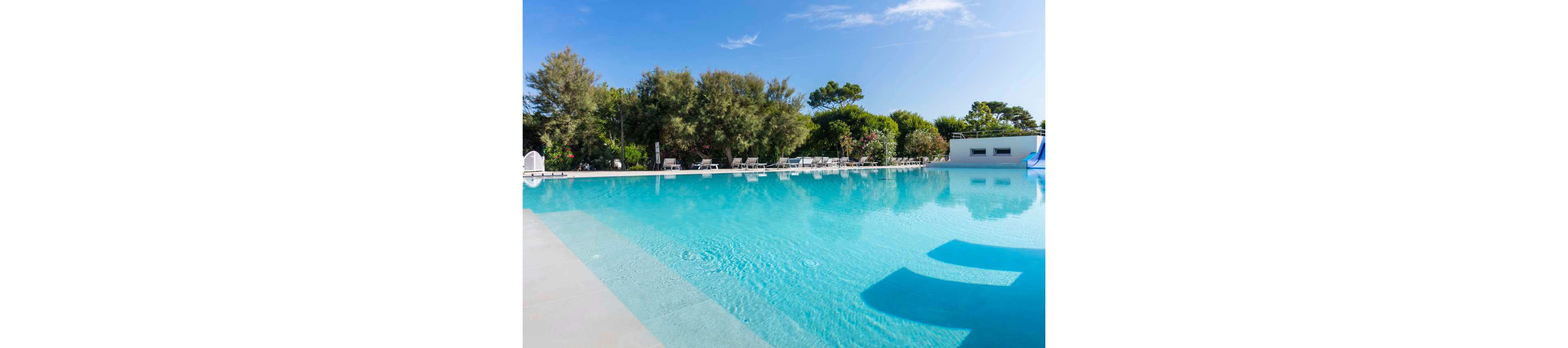 A large, clear swimming pool is featured outdoors under a bright blue sky. Lounge chairs line the poolside, surrounded by lush green trees and shrubs, creating a relaxing atmosphere. The water reflects the sky and nearby foliage, while a small white building and a blue slide are visible in the background. The setting is tranquil and inviting, ideal for leisure and relaxation.