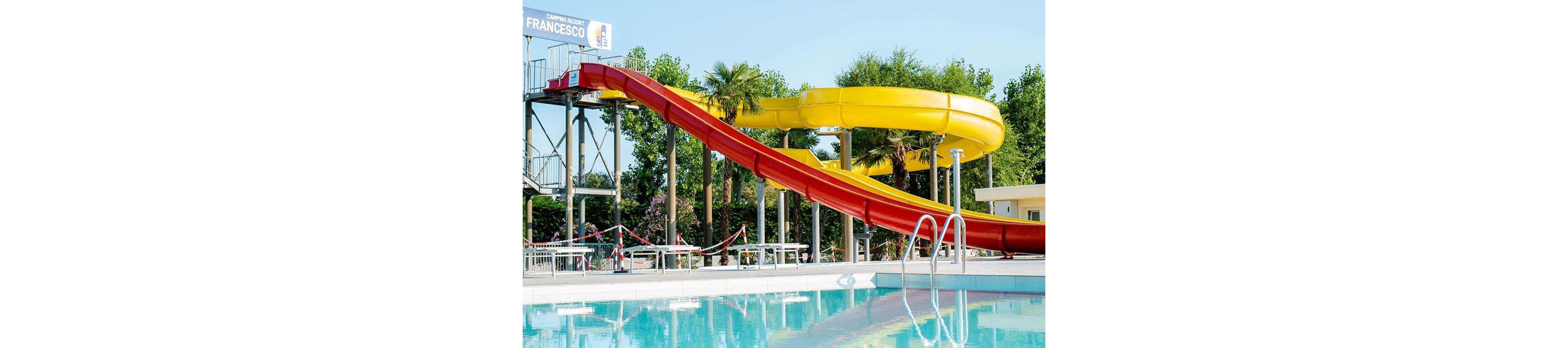 A large red and yellow water slide curves above a clear blue swimming pool in an outdoor recreational area. The setting features lush green trees and a sunny sky, with poolside seating and a visible sign reading 'FRANCESCO.' The scene is bright and inviting, emphasizing leisure and summer fun. No people are present in the image.