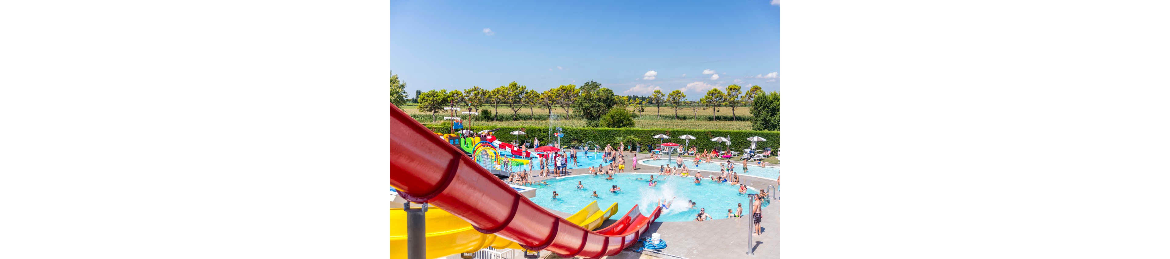 A lively outdoor water park features a large swimming pool filled with people of various ages enjoying the sunny day. Prominent red and yellow water slides curve into the pool, while children and adults play in the water and relax under white umbrellas. The background includes green trees, open fields, and a clear blue sky, creating a vibrant and energetic summer atmosphere. No visible text or numbers are present in the image.