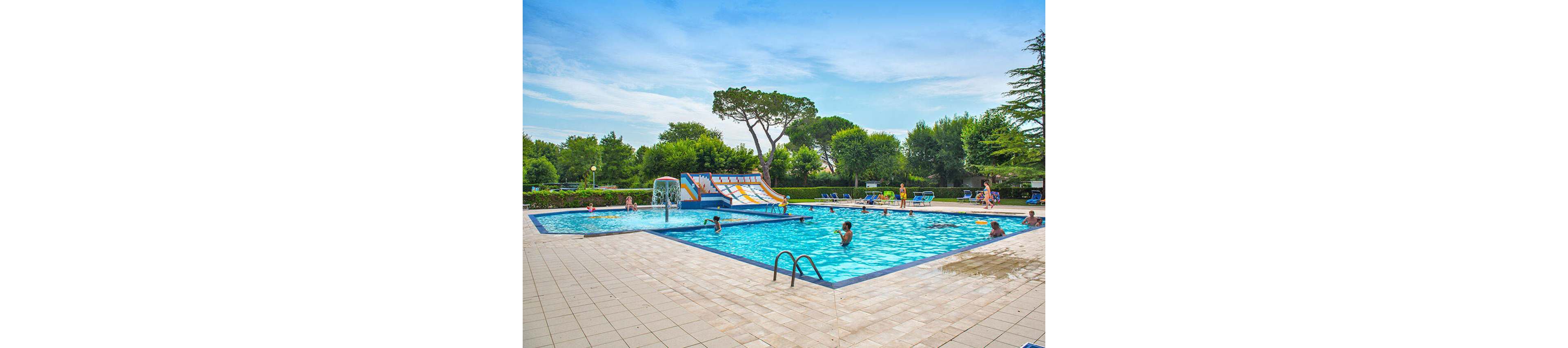 A large outdoor swimming pool features a colorful water slide and several children and adults enjoying the water. The pool is surrounded by a tiled deck, lounge chairs, and lush green trees under a bright blue sky. The atmosphere is lively and family-friendly, with people swimming, relaxing, and playing. No visible text or numbers are present in the image.