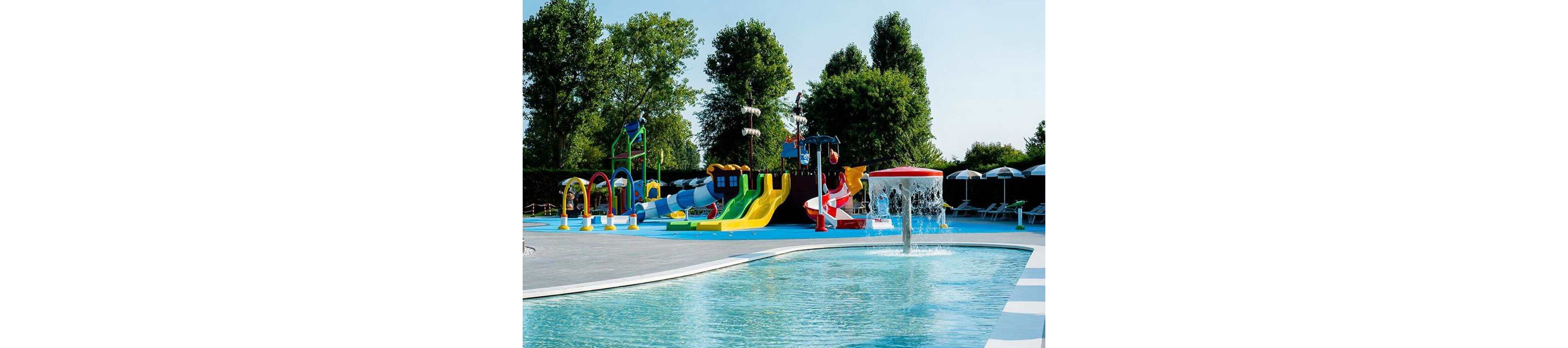 A vibrant outdoor water playground features multiple slides, splash structures, and interactive water elements. The setting is a sunny day with clear skies, surrounded by lush green trees and shaded seating areas. Brightly colored slides and water sprayers create a playful atmosphere, while a shallow pool with a central fountain invites children to play. The area is clean and well-maintained, ideal for family recreation and summer fun.