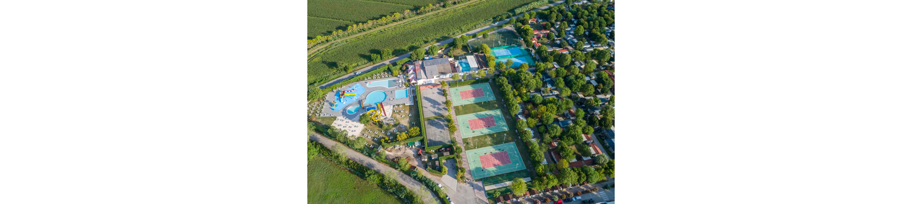 This aerial image captures a large recreational complex featuring multiple tennis courts, a water park with slides, and a swimming pool. The facility is surrounded by lush greenery and adjacent to a residential area. The courts have red and green surfaces, while the water park includes colorful slides and play structures. The setting is bright and vibrant, suggesting a lively and family-friendly atmosphere.