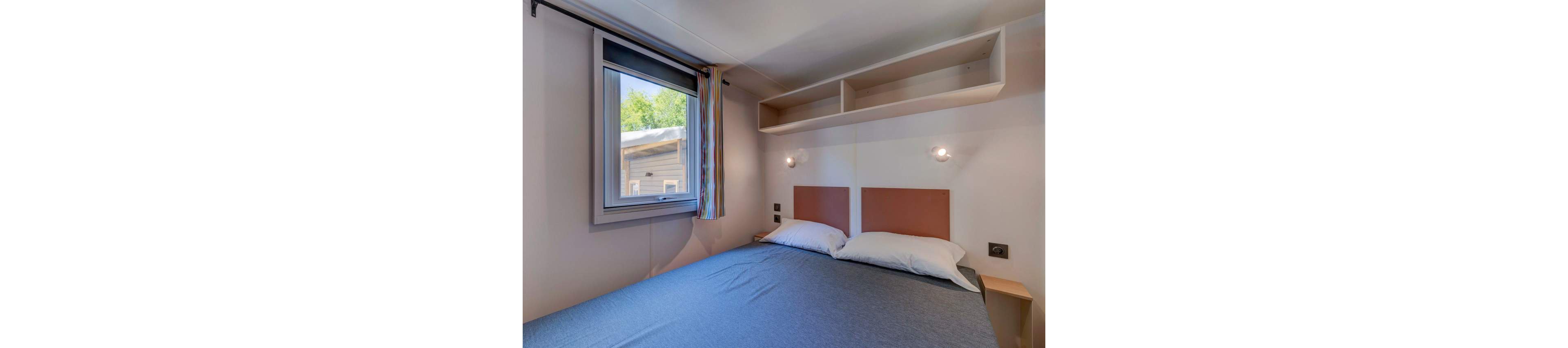 A small, modern bedroom features a double bed with two pillows and a blue-gray bedspread. The room includes a window with a view of greenery outside, colorful curtains, and built-in shelving above the bed. Two wall-mounted reading lights provide illumination, and the overall palette is neutral with soft natural light. No visible text or numeric values are present in the image.