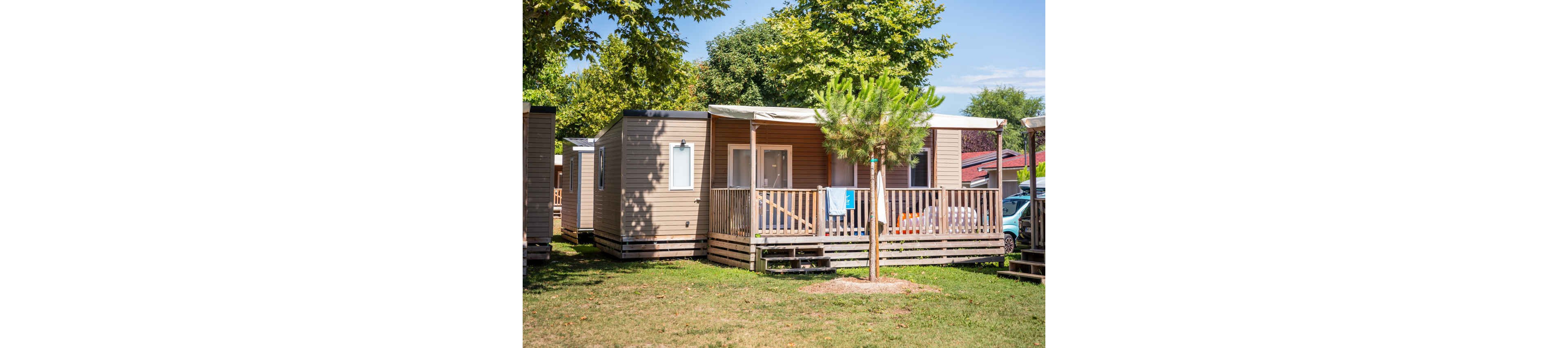 A contemporary mobile home is featured with a spacious wooden porch, set in a grassy area surrounded by trees. The scene is bright and sunny, evoking a relaxed vacation mood. A towel hangs on the porch railing, suggesting recent activity or occupancy. The mobile home is part of a cluster, with similar units visible nearby and a parked car partially seen in the background.