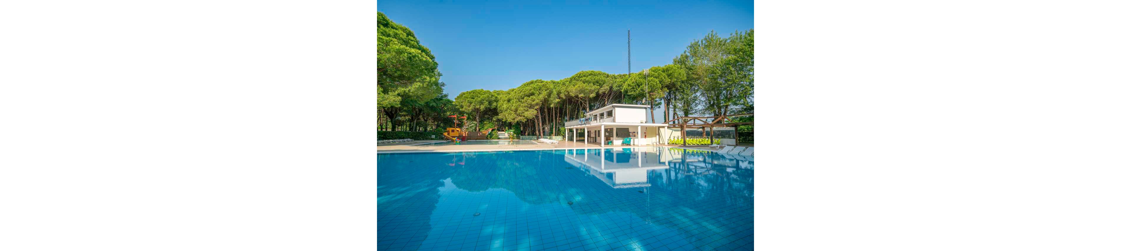 A large, clear blue swimming pool is the main focus, reflecting a modern white poolside building. The setting is outdoors, surrounded by lush green trees under a bright blue sky. In the background, a children's play area with water features is visible, adding a family-friendly atmosphere. The scene is calm and inviting, with no people present.