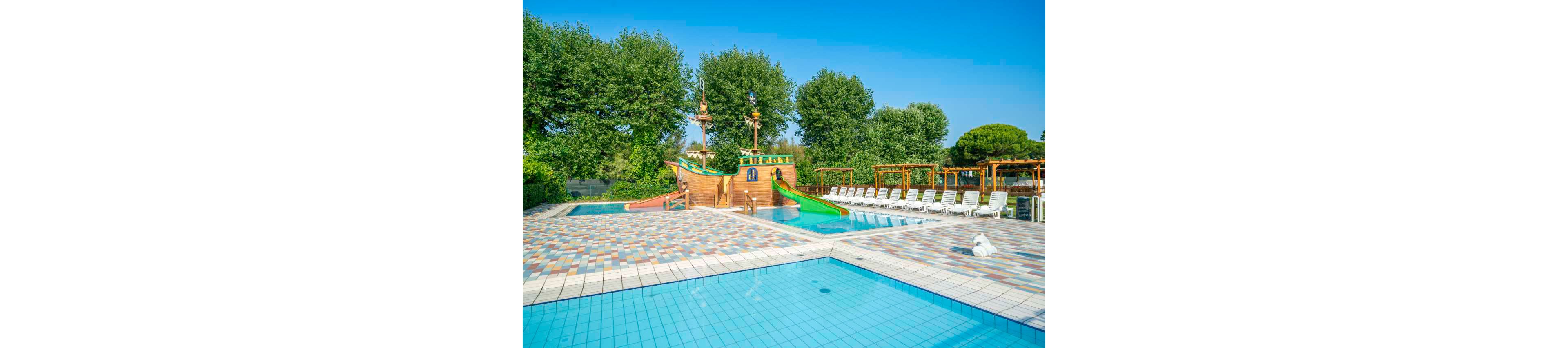 A vibrant outdoor swimming pool features a large pirate ship-themed play structure with colorful slides. The pool area is surrounded by lounge chairs and lush green trees, creating a family-friendly atmosphere. The tiled pool deck and clear blue water enhance the inviting setting. No visible people are present, and the sky is clear and sunny. No explicit numbers or text are visible in the image.