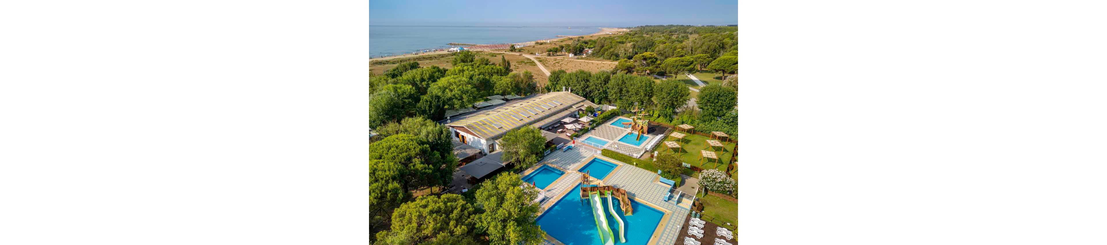 This image shows an outdoor water park complex with multiple swimming pools and water slides, surrounded by lush greenery. The pools feature colorful slides and play structures, and the facility is located close to a sandy beach visible in the background. The setting appears to be a family-friendly recreational area with shaded seating and open lawns. The overall mood is vibrant and inviting, ideal for summer activities. No visible text or numbers are present in the image.