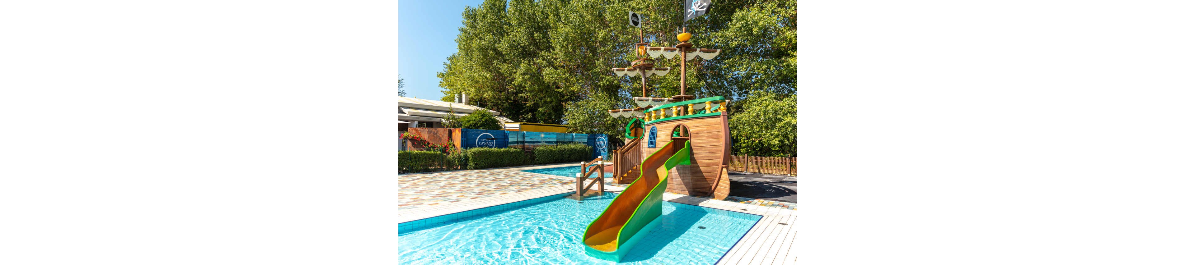 A group of children enjoy a sunny day at an outdoor pool featuring a large pirate ship-themed slide. The setting is a family-friendly water play area with clear blue water and playful visuals. The pirate ship structure includes a yellow and green slide, wooden details, and a black flag with a skull and crossbones. Lush green trees and a building are visible in the background, creating a vibrant and inviting atmosphere.