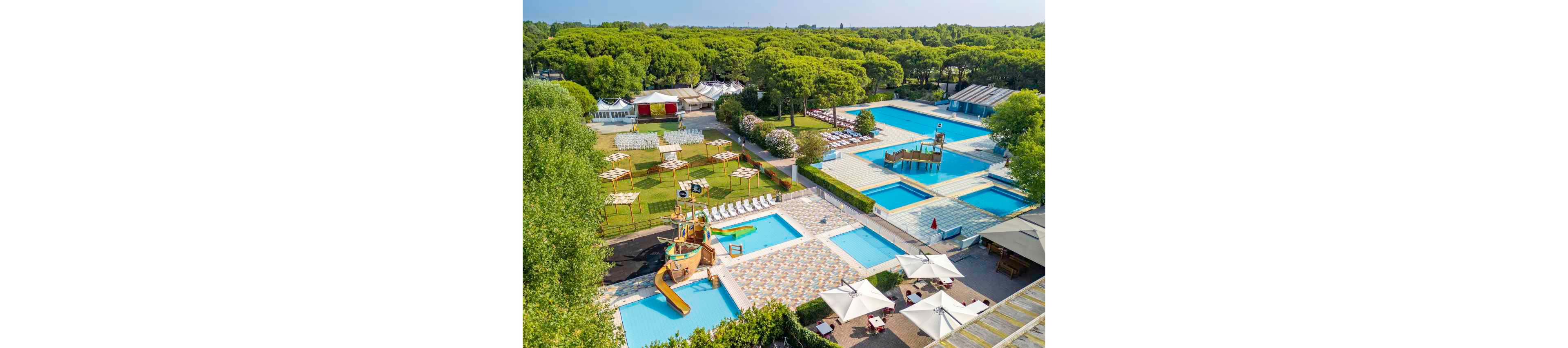This image shows an expansive outdoor water park with multiple swimming pools and a children's play area featuring slides and water features. The setting is surrounded by lush green trees and shaded seating areas with umbrellas. Several cabanas and tents are visible, suggesting a recreational or resort environment. The overall mood is vibrant and family-friendly, with a bright and sunny color palette. No visible text or numeric values are present in the image.