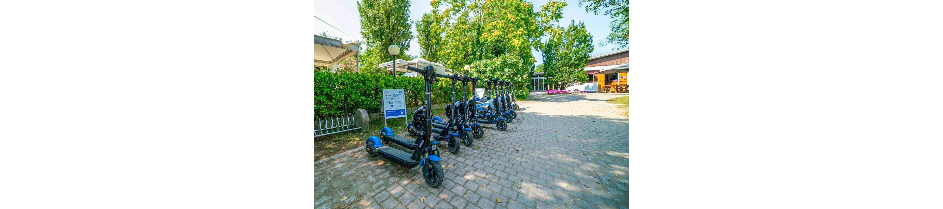 A line of electric scooters is neatly parked on a paved pathway in a leafy outdoor setting. The area is bright and sunny, with green trees and bushes surrounding the scooters. A sign with visible text stands next to the scooters, indicating information or instructions for users. The background includes a building and shaded seating area, suggesting a public or recreational space. The overall mood is vibrant and inviting, ideal for urban mobility.