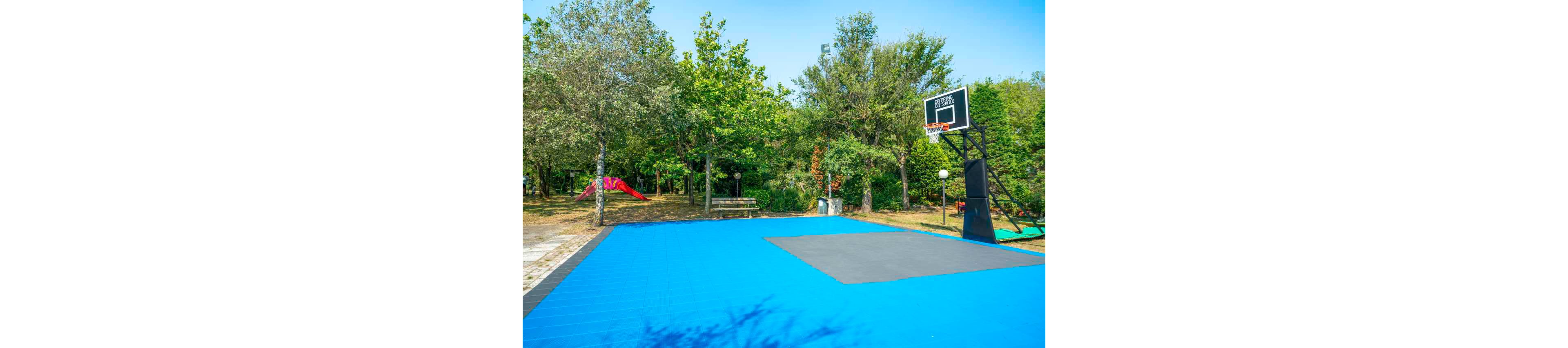 A vibrant blue and gray basketball court is set outdoors, surrounded by dense green trees and park amenities. The court features a single black basketball hoop with a visible backboard displaying the number '295'. In the background, a red playground slide and benches are visible, enhancing the recreational atmosphere. The scene is brightly lit, evoking a cheerful and inviting mood.
