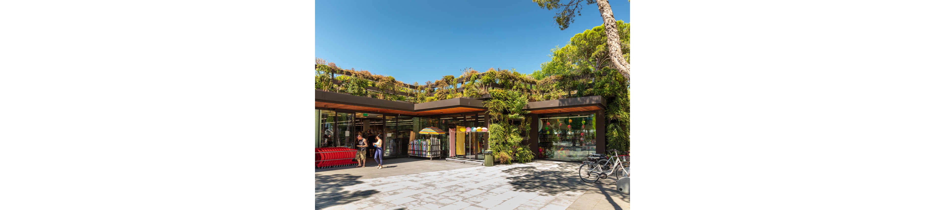 A contemporary retail store features a lush green roof and vertical gardens, blending architecture with nature. The entrance area displays colorful umbrellas and shopping carts, while bicycles are parked nearby. Two women stand outside, engaging in conversation under a sunny sky. The scene conveys a vibrant, sustainable, and welcoming atmosphere.