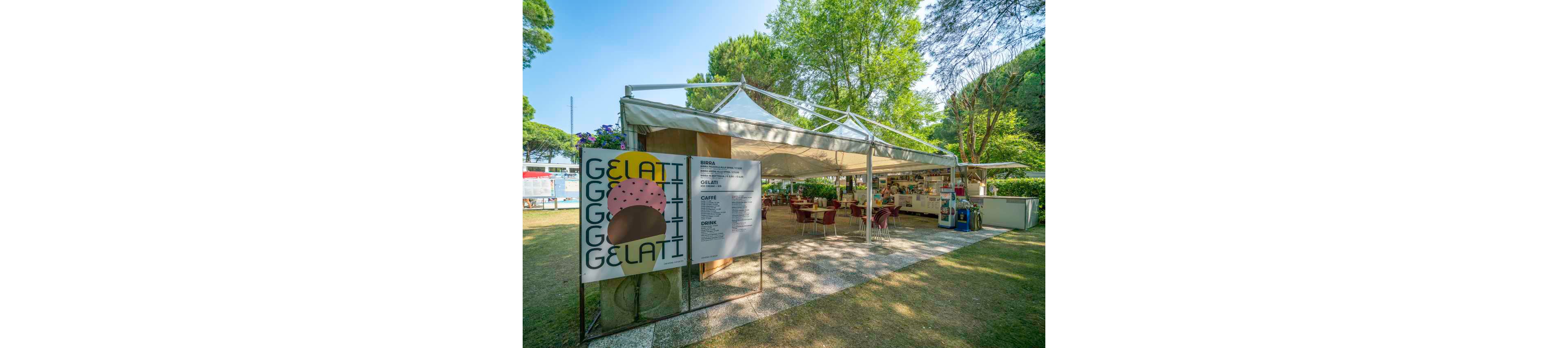 An open-air café features a large sign with the word 'GELATI' and an illustrated ice cream cone, inviting visitors to enjoy frozen treats. The setting is a shaded garden area with trees, outdoor tables, and a white canopy tent. The menu board is visible, listing various gelato and coffee options. The atmosphere is relaxed and summery, with natural light and greenery surrounding the café. No specific brands or numeric values are clearly visible.