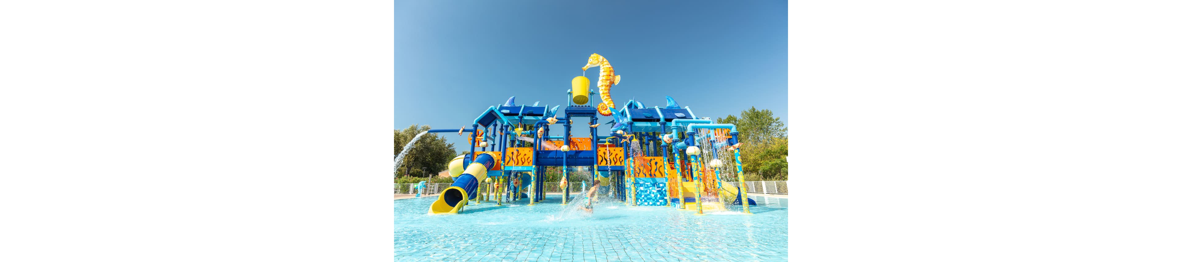 A vibrant water playground features multiple slides, climbing structures, and a large tipping bucket under a clear blue sky. Children are actively playing and splashing in the shallow pool area, surrounded by playful aquatic-themed decorations. The scene is bright and cheerful, with a dominant palette of blue, yellow, and orange, and lush greenery in the background.
