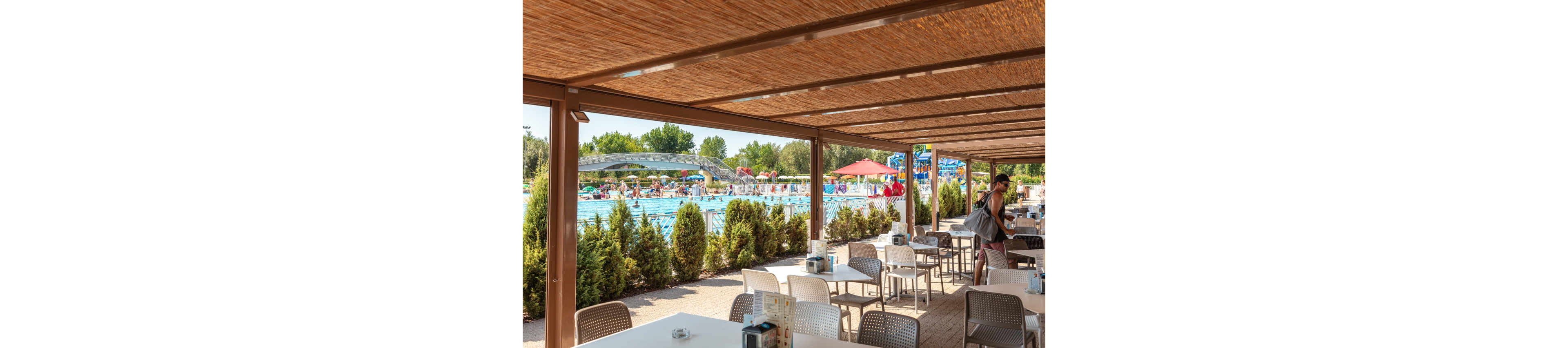 A shaded outdoor café area overlooks a busy swimming pool filled with people enjoying summer activities. The seating area features modern tables and chairs under a wooden pergola, with lush greenery providing separation from the pool. In the background, swimmers, sunbathers, and colorful umbrellas create a lively, relaxed atmosphere. A man with a backpack walks through the café, adding to the casual vacation vibe.