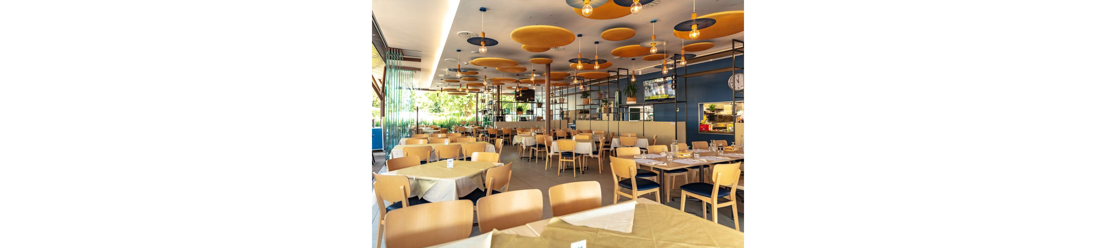 A spacious, contemporary restaurant dining area features light wooden chairs and tables set with white and beige linens. The ceiling is decorated with circular yellow and blue acoustic panels, and pendant lights add a warm ambiance. Large windows allow natural light to fill the space, while greenery is visible outside. The setting is clean, inviting, and designed for casual dining experiences.
