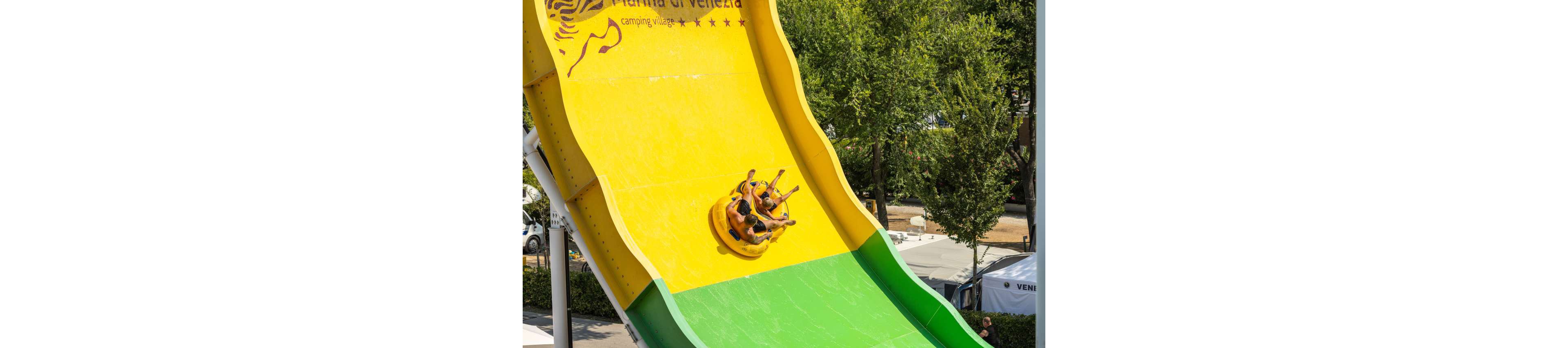 A group of people is riding an inflatable raft down a steep, brightly colored water slide. The slide features bold yellow and green sections and is located outdoors, surrounded by trees and tents. The visible text at the top reads 'Praia di Velezia camping village' with a stylized zebra graphic. The scene captures a fun, energetic moment on a sunny day.