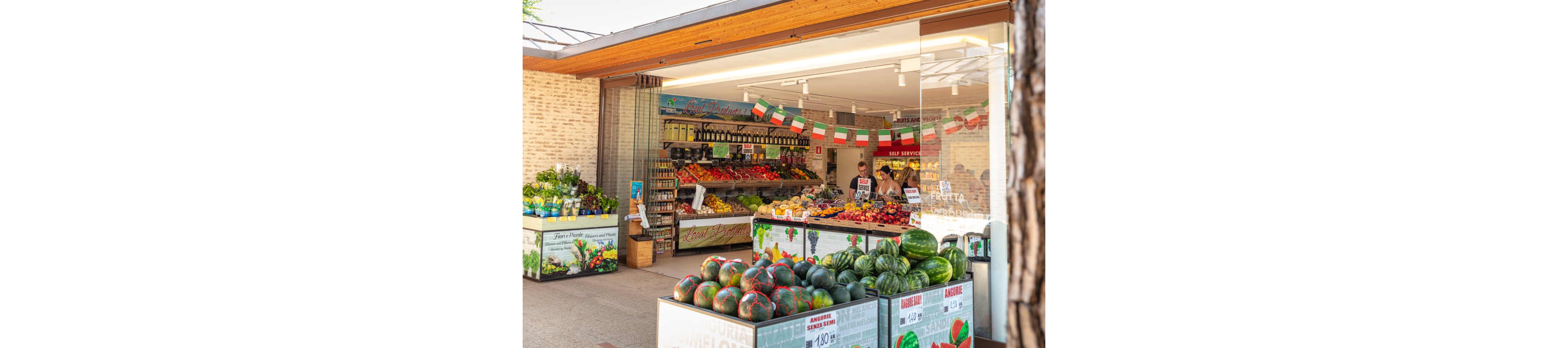 A vibrant outdoor market stall showcases a variety of fresh fruits and vegetables, including watermelons, tomatoes, and citrus. The setting is a modern, open-air shop with Italian flags and signage, suggesting a festive or local atmosphere. Several people, including adults, are seen shopping and working behind the counter. Visible numeric values such as '4,10' and '10,99' are displayed on price tags. The overall mood is lively and colorful, with a focus on freshness and abundance.