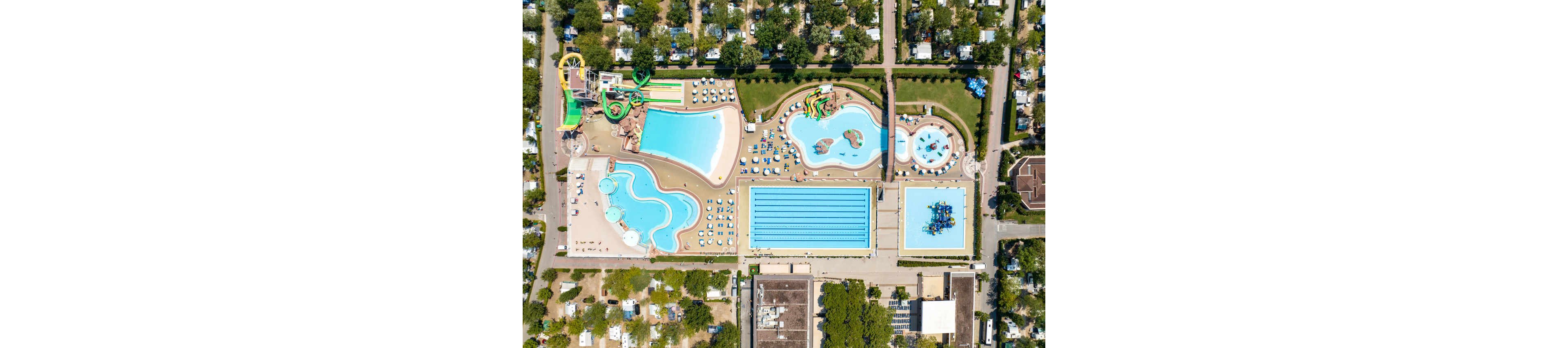 A vibrant aerial photograph captures a sprawling outdoor waterpark featuring multiple swimming pools, water slides, and play areas. The scene includes a large lap pool with visible lane markings, several irregularly shaped leisure pools, and colorful water slides. Sun loungers and umbrellas are arranged around the pools, and the surrounding area is filled with trees and buildings, suggesting a resort or campground setting. The image is bright and lively, with a clear, sunny atmosphere.