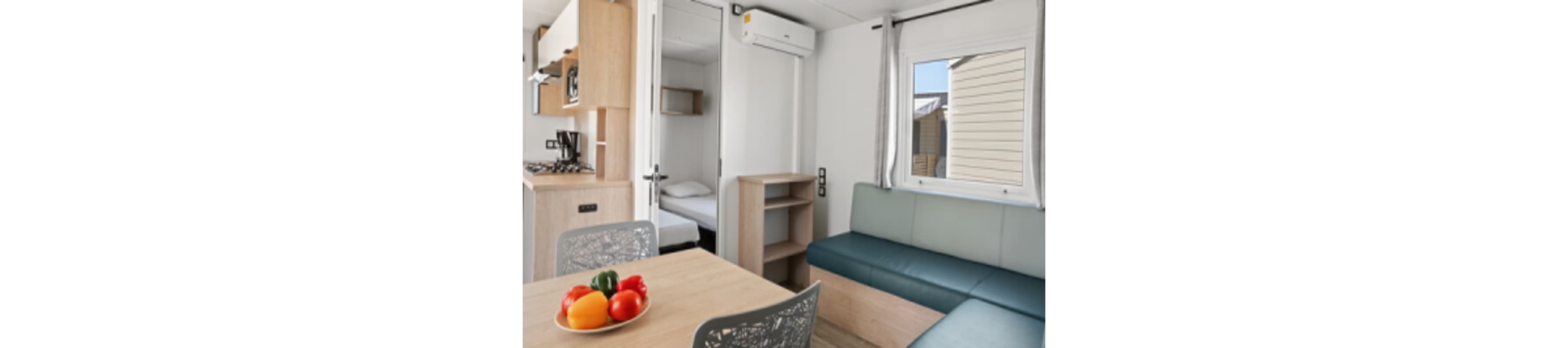 A bright, modern mobile home interior features a small kitchen with a stovetop and microwave, a dining table with colorful bell peppers, and a cozy seating area. The space is decorated in light wood tones and soft blue-green upholstery, with a visible air conditioning unit above the doorway. Natural light enters through a window, and a small bedroom with a single bed is visible in the background.