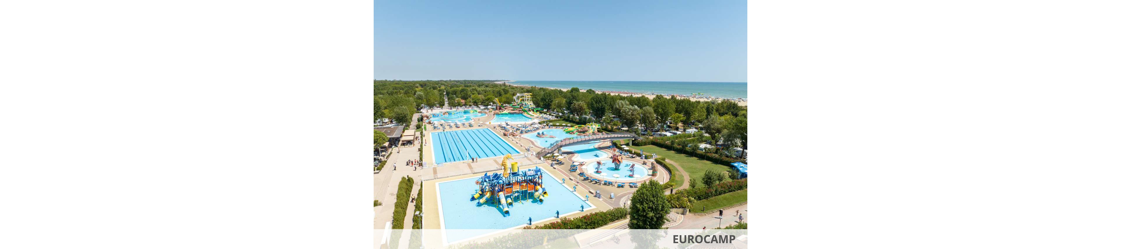 This image shows a large outdoor water park complex with multiple swimming pools, water slides, and play areas. The setting is adjacent to a sandy beach and surrounded by lush greenery and trees. The pools include a lap pool, a children's splash zone with colorful structures, and circular pools, all under a clear blue sky. People of various ages are visible enjoying the facilities, and the overall mood is bright and lively.