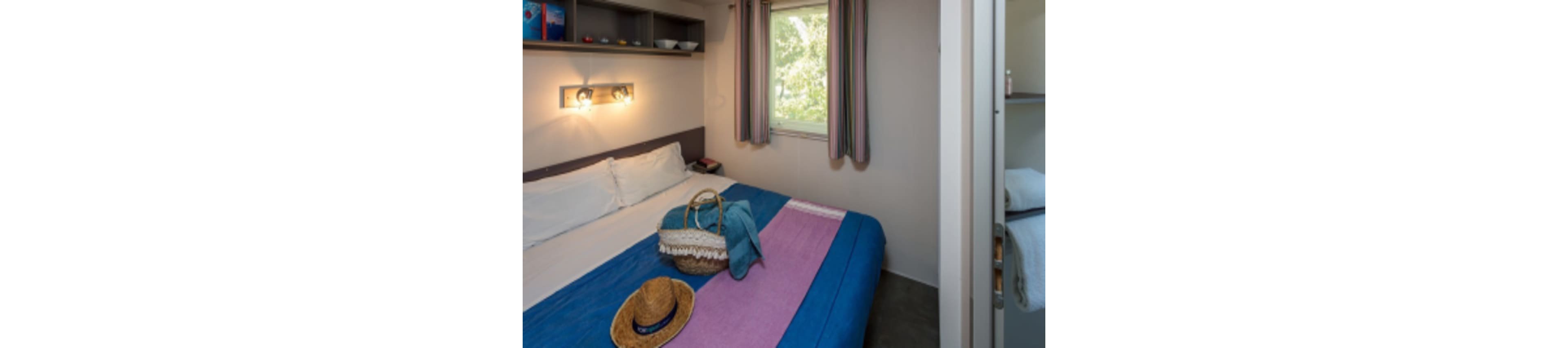 A neatly made double bed with a blue and purple blanket is the main focus of this small, cozy bedroom. Natural light enters through a window with striped curtains, and a straw hat and woven bag rest on the bed, suggesting a relaxed, vacation atmosphere. Shelves above the bed hold books and small items, while a soft wall lamp adds warmth to the scene. The overall palette is bright and inviting, with hints of outdoor greenery visible.