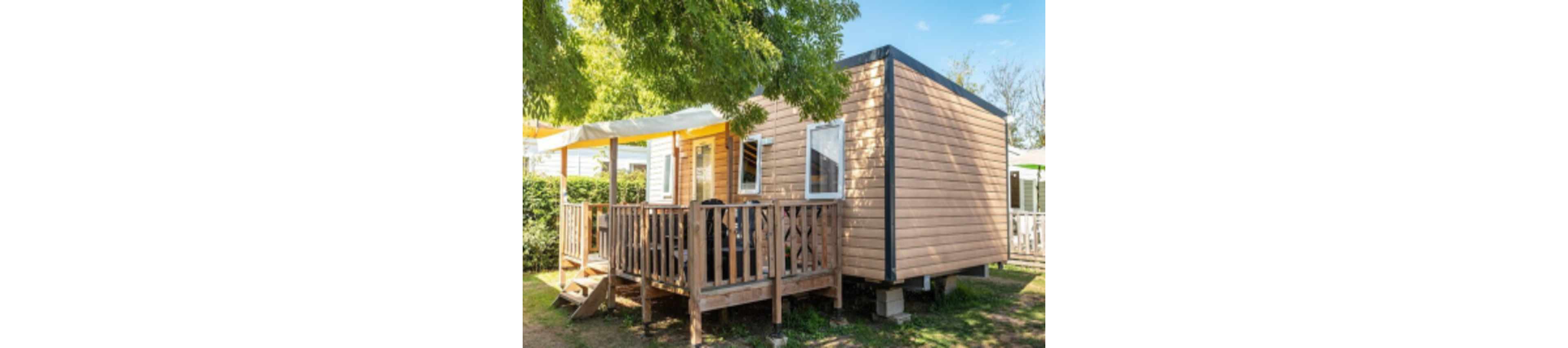 A contemporary wooden mobile home is shown with a covered porch and railing, set outdoors under leafy green trees. The structure features large windows and a shaded seating area, creating a welcoming and relaxed atmosphere. Sunlight filters through the trees, highlighting the natural wood texture and peaceful garden setting. No visible text or numbers are present in the image.