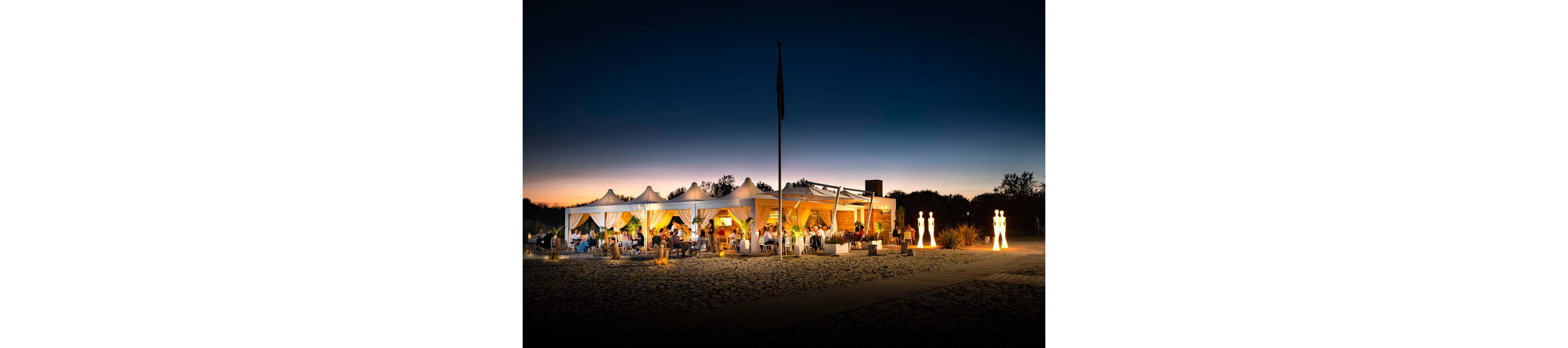 A stylish beachfront restaurant is illuminated at dusk, with guests dining under white canopy tents. The setting features soft, ambient lighting and a sandy beach foreground, creating a relaxed yet sophisticated atmosphere. The sky transitions from deep blue to warm hues, enhancing the tranquil mood. Several decorative statues are visible near the entrance, adding to the upscale ambiance.