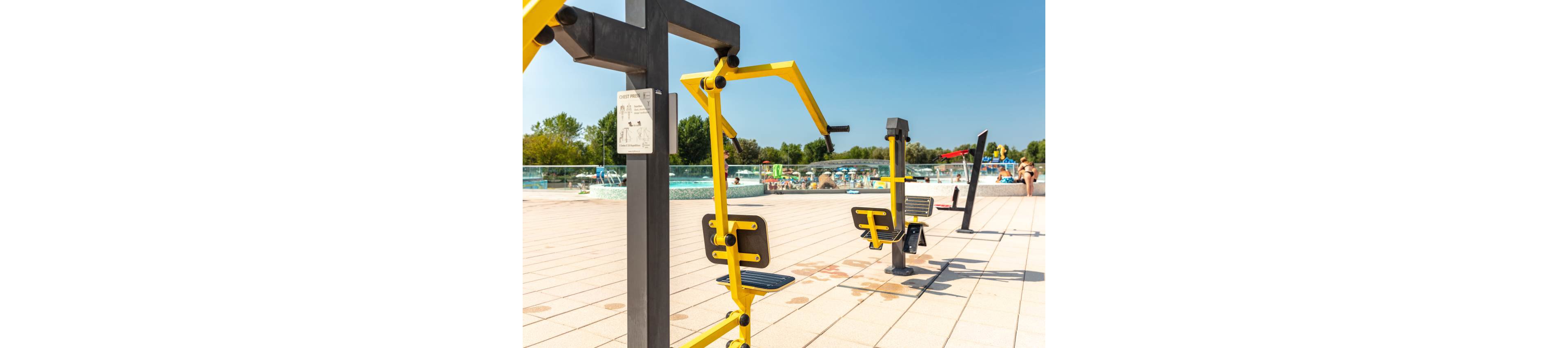 Bright yellow and black outdoor gym machines are set up on a spacious tiled patio beside a large swimming pool. The scene is sunlit with clear blue skies, and people are visible relaxing by the pool in the background. The equipment is designed for public use, with instructional signage attached. The overall mood is active and inviting, with a modern recreational setting.