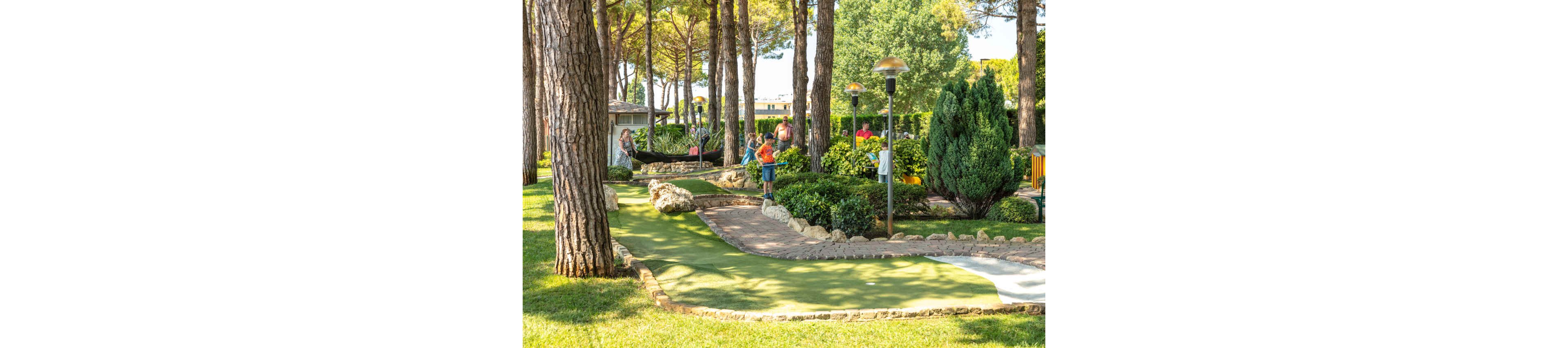 A group of people, including children and adults, play mini golf on a well-maintained outdoor course surrounded by lush greenery and tall trees. The setting is bright and sunny, with landscaped gardens and decorative rocks enhancing the scene. Lamp posts and benches are visible, adding to the park-like atmosphere. The mood is relaxed and cheerful, with families engaging in a fun recreational activity. No visible text or numbers are present in the image.