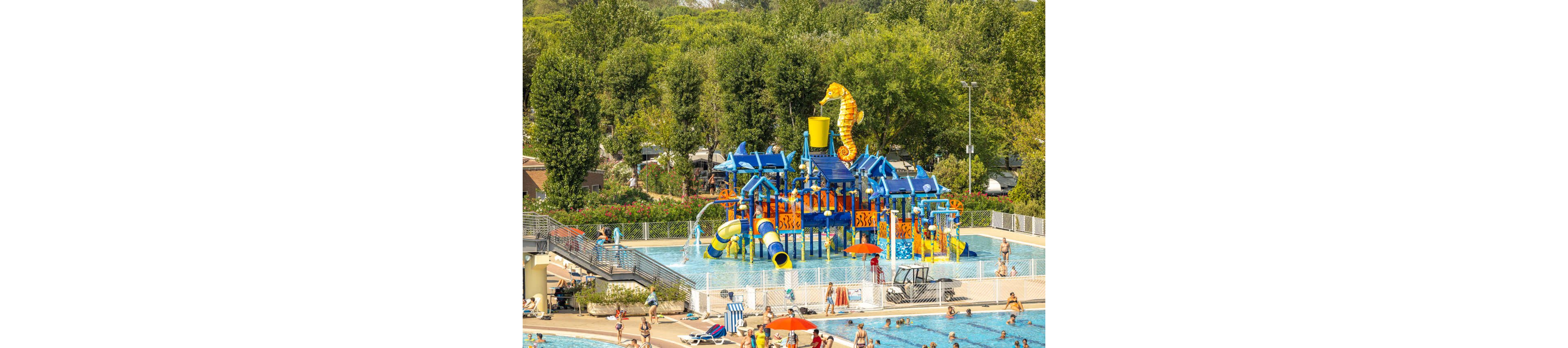 A vibrant water playground featuring a large seahorse slide stands in the center of a public swimming pool. Children and adults enjoy various water activities, with slides, splash features, and a giant tipping bucket. The setting is outdoors, surrounded by lush green trees and a sunny atmosphere. The pool area is busy with families and groups, creating a lively and playful mood.