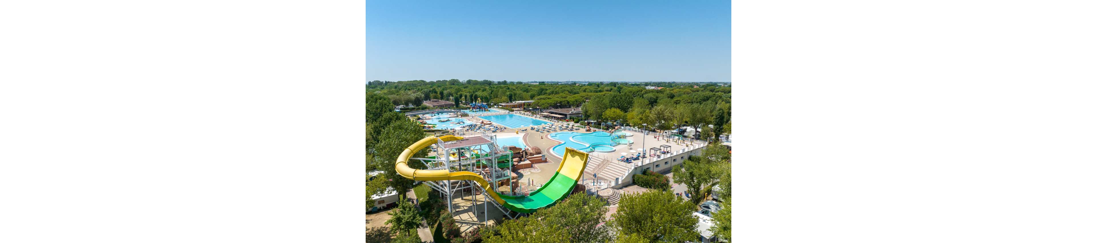 Aerial view of a spacious outdoor water park featuring several swimming pools and a prominent yellow and green water slide. The setting is surrounded by lush greenery and trees, with clear blue skies overhead. The park includes lounging areas, sunbeds, and various pool shapes, creating a lively and inviting atmosphere. No visible text or numbers are present in the image.