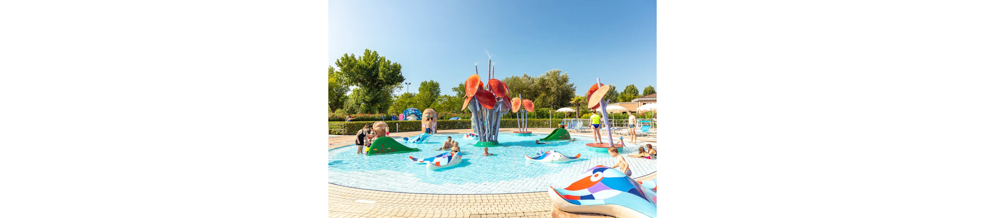 A group of children and adults enjoy a vibrant outdoor splash pool featuring whimsical animal sculptures and large flower-shaped water features. The setting is a sunny day at a family-friendly water park, with clear blue skies and lush greenery in the background. The pool area is designed for young children, with shallow water and playful, interactive elements. No visible text or numbers are present in the image.