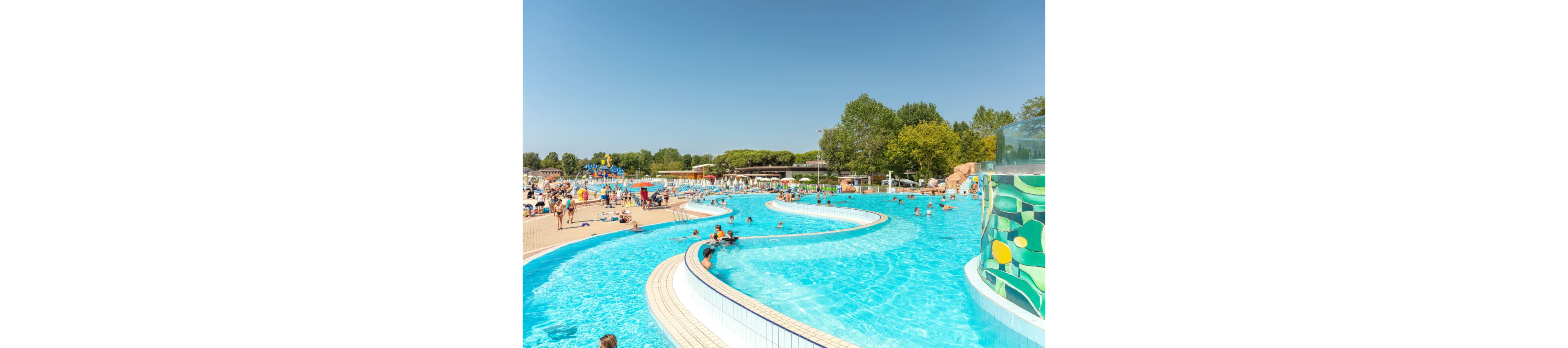 A large outdoor water park features a winding lazy river pool filled with people enjoying a sunny day. The setting includes families and children swimming and relaxing, with lush green trees and clear blue skies in the background. The pool area is surrounded by sunbathers, colorful slides, and recreational facilities. The overall mood is lively and summery, with bright blue water and vibrant visuals. No visible text or numbers are present in the image.