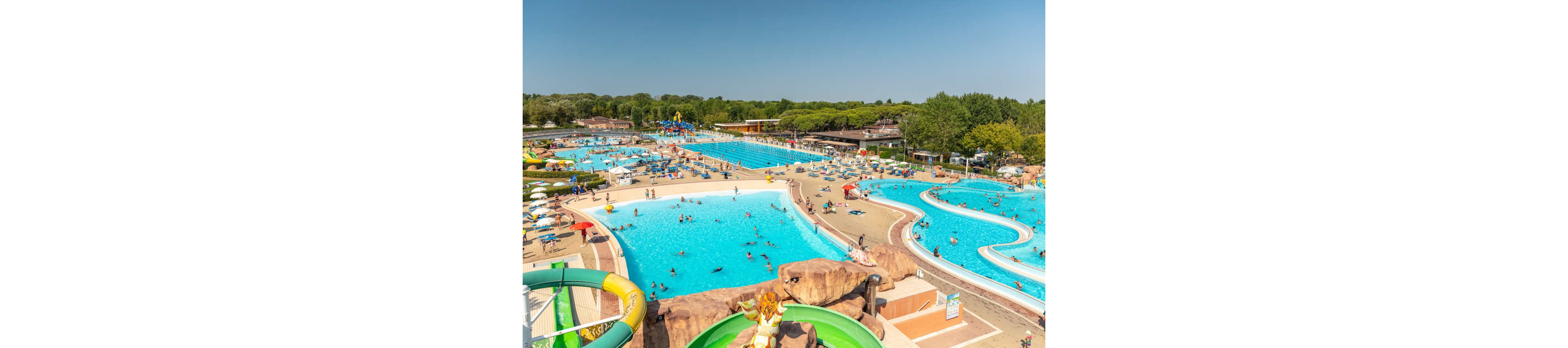 A sprawling outdoor waterpark features several large swimming pools and winding water slides under a clear blue sky. Families and children enjoy swimming and playing in the water, while others relax on the poolside deck. The setting is lively and vibrant, with lush green trees in the background and a summery atmosphere. The pools are surrounded by walkways and sunbathing areas, creating a fun and energetic environment. No visible text or numeric values are present in the image.
