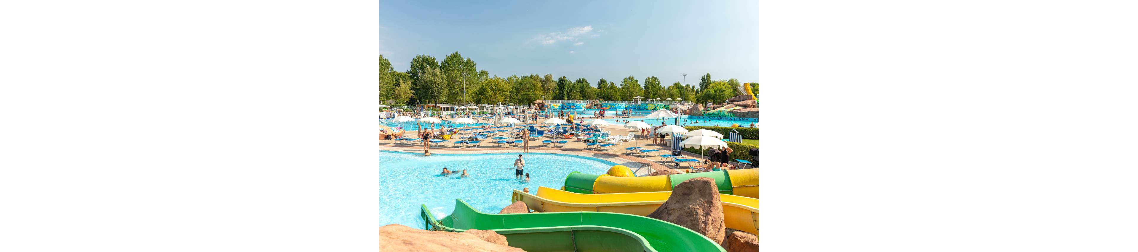A lively outdoor water park features multiple pools, colorful water slides, and numerous sun loungers under umbrellas. Families and children enjoy swimming and relaxing in the bright, sunny weather. The setting is surrounded by green trees, creating a vibrant and inviting summer atmosphere. No visible text or numbers are present in the image.
