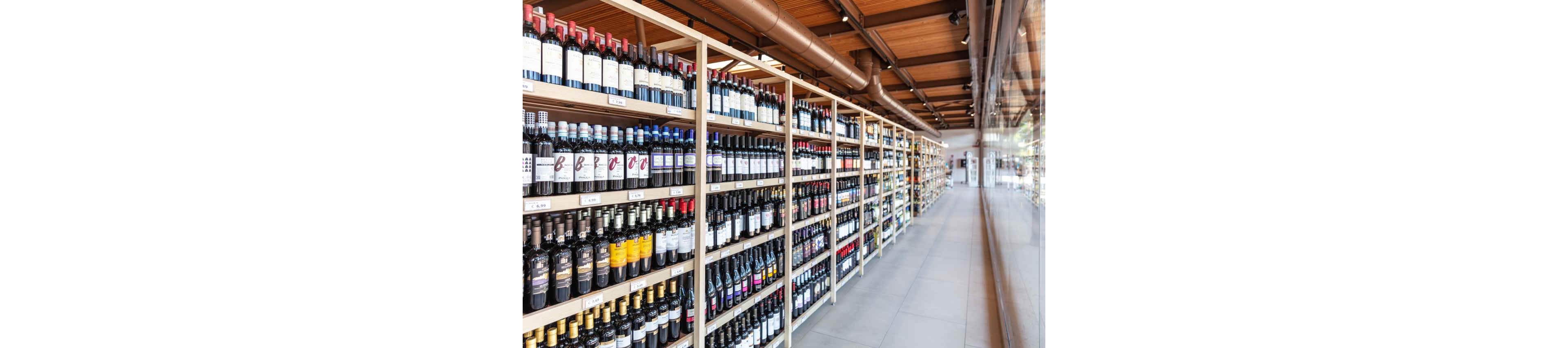 A spacious wine shop features multiple rows of wooden shelves filled with assorted wine bottles. The setting is bright and contemporary, with large windows reflecting the organized display. The image highlights the variety of wine labels and bottle shapes, with no visible people present. The overall mood is clean and inviting, emphasizing abundance and selection.